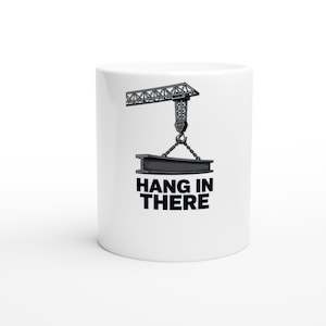 May include: A white ceramic mug featuring a black graphic illustration of a construction crane lifting an I-beam with chains. Below the illustration, the text HANG IN THERE is printed in bold, black, capital letters on the front of the mug.