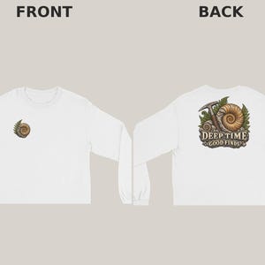 May include: A white long-sleeve shirt featuring a small ammonite fossil graphic on the front left chest. The back displays a larger illustration of an ammonite fossil, a rock hammer, and fern leaves with the text Deep Time Good Finds.