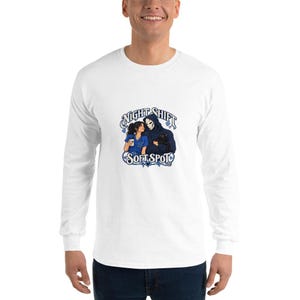 Op de afbeelding: Een wit shirt met lange mouwen en een grafische print van een verpleegkundige in blauwe kleding met een koffiekop naast een figuur met een capuchon in een donkerblauwe mantel die een zwarte kat vasthoudt. De tekst luidt Night Shift Soft Spot.