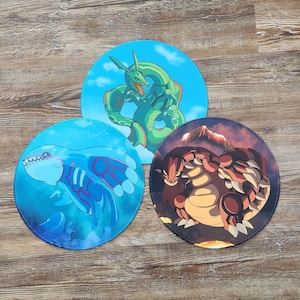 May include: Three round, colorful mousepads featuring different Pokemon characters. The first mousepad is blue with a light blue Pokemon with red accents. The second mousepad is green with a green Pokemon with red accents. The third mousepad is brown with a brown Pokemon with red accents.