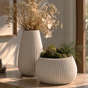 May include: A tall, white, ribbed cylindrical vase filled with dried baby's breath and a shorter, white, ribbed, rounded bowl-shaped planter containing green moss and small fern-like sprigs, both featuring a consistent vertical texture.