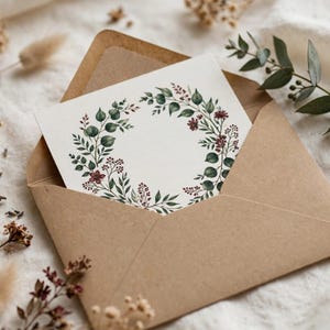 May include: A white rectangular card featuring a circular watercolor illustration of green eucalyptus leaves and small dark red berries, tucked inside a brown kraft paper envelope. The design forms a wreath shape with an empty center space.