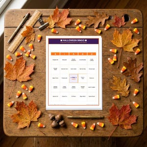 May include: A Halloween Bingo game card featuring a 5x5 grid with text labels like Pumpkin Pie, Flying Bat, and Ghostly Footprints. The card has a purple header and orange grid lines, surrounded by scattered autumn leaves and candy corn pieces.