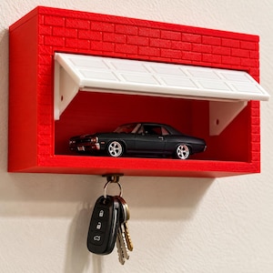 May include: A red wall-mounted key holder shaped like a brick garage with a white overhead door. Inside the garage sits a black toy muscle car. A metal ring with several keys hangs from a hook attached to the bottom of the garage structure.