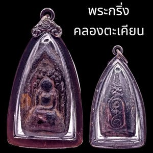 May include: Two dark, textured, metallic amulets encased in silver-toned frames with loops for hanging. The larger amulet features a seated figure, while the smaller one displays intricate engraved symbols. Thai text reads พระกริ่ง คลองตะเคียน.