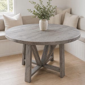 Op de afbeelding: Een ronde eettafel met een verweerde grijze houten afwerking en een stevig onderstel. Een crèmekleurige keramische vaas met groene eucalyptus takken staat in het midden van het tafelblad, dat een zichtbare houtnerfstructuur heeft.