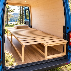 Pode incluir: Uma estrutura de cama de ripas de madeira clara instalada dentro da área de carga traseira de uma carrinha azul. A estrutura apresenta uma base retangular com ripas horizontais e um cobertor bege dobrado sobre a superfície.