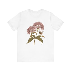 Op de afbeelding: Een wit t-shirt met korte mouwen voorzien van een gecentreerde botanische illustratie van een roze bloementros met groene bladeren en een hommel die bij de bloesems zweeft. De afbeelding is gedetailleerd met gedempte tinten en vormen.