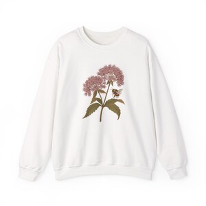 Op de afbeelding: Een wit sweatshirt met lange mouwen en een centrale grafische illustratie van een roze bloeiende plant met groene bladeren en een kleine hommel die bij de bloesems vliegt. Het kledingstuk heeft een ronde hals en geribbelde boorden.
