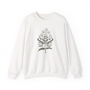 May include: A white long-sleeve crewneck sweatshirt featuring a central graphic illustration of a moth perched on a flowering plant. The moth has detailed black and white wings, while the plant has green leaves and clusters of small white flowers.