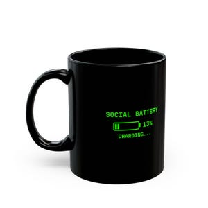 Op de afbeelding: Een zwarte keramische mok met een felgroene afbeelding van een batterijpictogram met het label SOCIAL BATTERY, dat een oplaadniveau van 13% aangeeft met de tekst CHARGING... in een digitaal lettertype onder het pictogram.