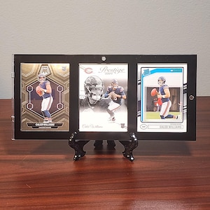 May include: A black triple-card display frame holding three Caleb Williams football trading cards. The left card is a gold Mosaic rookie card, the center is a white Prestige card, and the right is a white Donruss card. The frame stands on a black easel.
