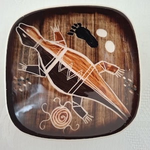 Puede incluir: Un plato cuadrado de cerámica con esquinas redondeadas y un fondo de color marrón oscuro con vetas de madera. El centro muestra una ilustración estilizada de un lagarto en tonos marrones y negros con contornos blancos, una huella negra, dos óvalos blancos y una espiral.