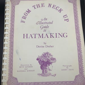Puede incluir: Un libro de instrucciones con encuadernación en espiral titulado From the Neck Up: An Illustrated Guide to Hatmaking de Denise Dreher. La portada es de color blanco roto con texto morado y un borde decorativo, con una ilustración de perfil de una persona con sombrero.