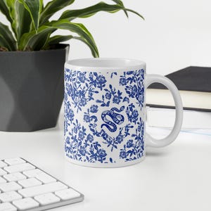 Può includere: Tazza in ceramica bianca con un motivo floreale blu e un'illustrazione di un serpente blu. La tazza ha un manico bianco. Un vaso di fiori e una tastiera sono sullo sfondo. La tazza è su una superficie bianca.