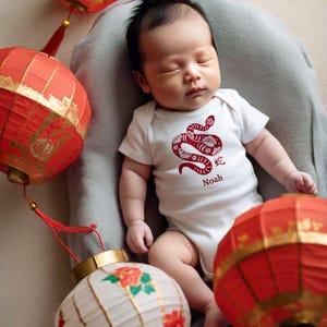 Peut inclure: Un body blanc pour bébé avec un motif de serpent rouge et le caractère chinois pour serpent. Le body porte également le nom "Noah" imprimé dessus. Le bébé est entouré de lanternes chinoises rouges.