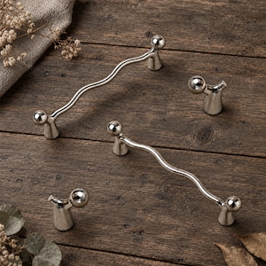 Peut inclure: Deux poignées de tiroir en métal argenté au design ondulé et allongé, accompagnées de deux boutons assortis en métal argenté, chacun doté d'un sommet sphérique et d'une base évasée, disposés sur une surface en bois sombre.