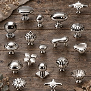 Könnte beinhalten: Eine Sammlung verschiedener polierter, silberfarbener Metall-Schrankknöpfe und Griffe in diversen Formen, darunter kugelförmige, geriffelte, florale und geometrische Designs. Jedes Stück hat eine reflektierende, glatte Oberfläche.