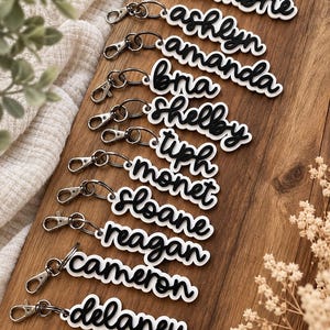 May include: A collection of ten custom keychains featuring black cursive names layered over white backing plates. Each keychain includes a silver-toned metal lobster clasp. The names displayed are cambre, ashlyn, amanda, bria, shelby, tiph, monet, sloane, reagan, cameron, and delaney.