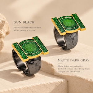 May include: Two rings featuring a green circular emblem with a gold border. One ring has a smooth, reflective gun black band, while the other has a matte dark gray band with a textured surface. Both rings display a green, granular inlay.