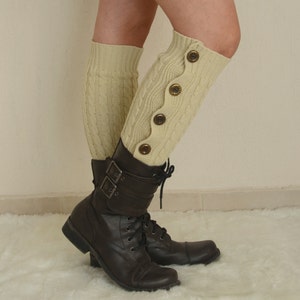 May include: A pair of cream-colored knitted leg warmers with buttons on one side. The leg warmers are worn with brown leather boots.