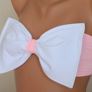 Può includere: Un top bandeau bianco e rosa con un grande fiocco sul davanti. Il fiocco è fatto di tessuto bianco con un centro rosa.