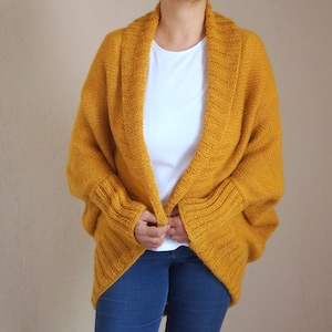 Puede incluir: Un c&aacute;rdigan de punto amarillo mostaza con cuello ancho y abierto y mangas largas. El c&aacute;rdigan est&aacute; drapeado sobre los hombros y tiene un corte holgado.