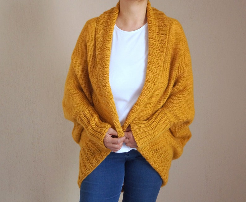 Puede incluir: Un c&aacute;rdigan de punto amarillo mostaza con cuello ancho y abierto y mangas largas. El c&aacute;rdigan est&aacute; drapeado sobre los hombros y tiene un corte holgado.