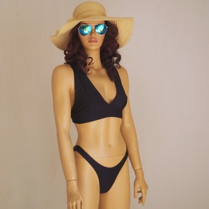 Può includere: Un top e un fondo bikini neri con una profonda scollatura a V. La modella indossa un cappello di paglia con occhiali da sole a specchio tinti di blu.