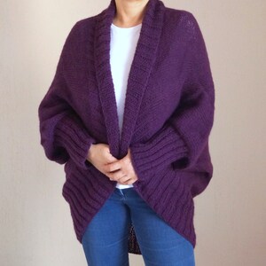 May include: A deep purple, open-front cardigan with a shawl collar. The knitted sweater has ribbed detailing on the sleeves and a relaxed fit. The person is wearing blue jeans and a white t-shirt underneath.