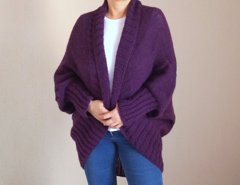 May include: A deep purple, open-front cardigan with a shawl collar. The knitted sweater has ribbed detailing on the sleeves and a relaxed fit. The person is wearing blue jeans and a white t-shirt underneath.