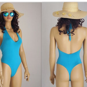 Puede incluir: Un traje de baño turquesa de estilo halter con un escote en V profundo. El traje de baño tiene una pierna de corte alto y una espalda de tanga.