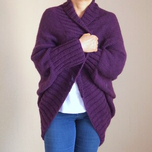 May include: A deep purple, open-front cardigan sweater. The sweater features a wide shawl collar and ribbed cuffs. The knitted design is visible, and the sweater appears to be made of a soft, warm material. The person is wearing blue jeans and a white shirt.