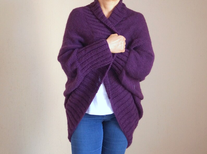 May include: A deep purple, open-front cardigan sweater. The sweater features a wide shawl collar and ribbed cuffs. The knitted design is visible, and the sweater appears to be made of a soft, warm material. The person is wearing blue jeans and a white shirt.
