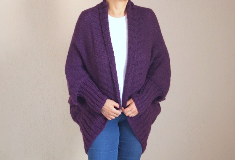 May include: A deep purple, open-front cardigan sweater with a shawl collar and ribbed cuffs. The sweater is made of a thick knit material and has a relaxed fit. The sleeves are long and the body is mid-length.