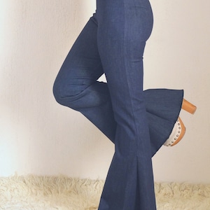 May include: Dark blue denim flare pants. The pants have a high waist and a wide bell bottom. The model is wearing white shoes and a white top. The pants are made of a soft denim material.