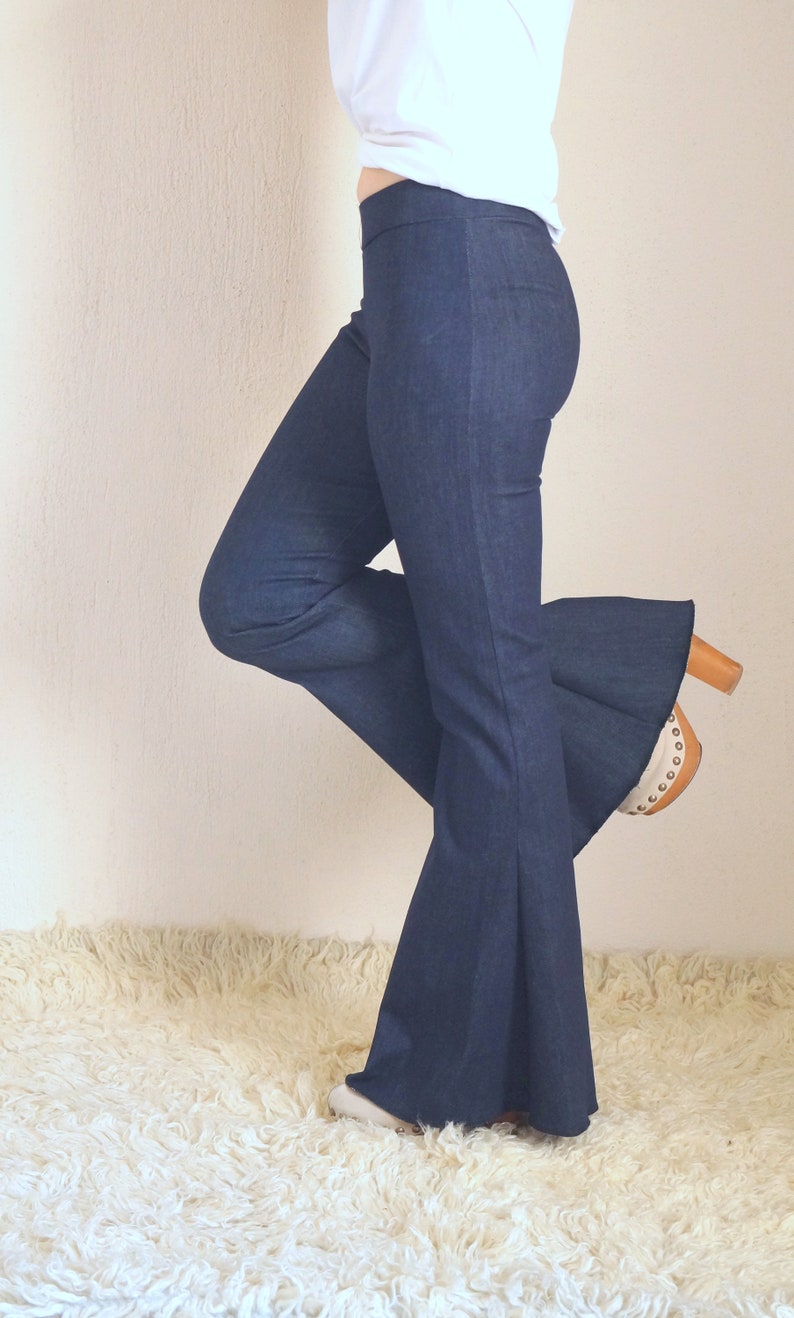 May include: Dark blue denim flare pants. The pants have a high waist and a wide bell bottom. The model is wearing white shoes and a white top. The pants are made of a soft denim material.