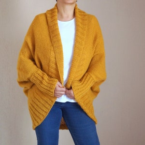 Puede incluir: Un c&aacute;rdigan de punto amarillo mostaza con cuello amplio abierto y mangas largas. El c&aacute;rdigan est&aacute; hecho de un hilo suave y grueso y tiene un corte holgado.