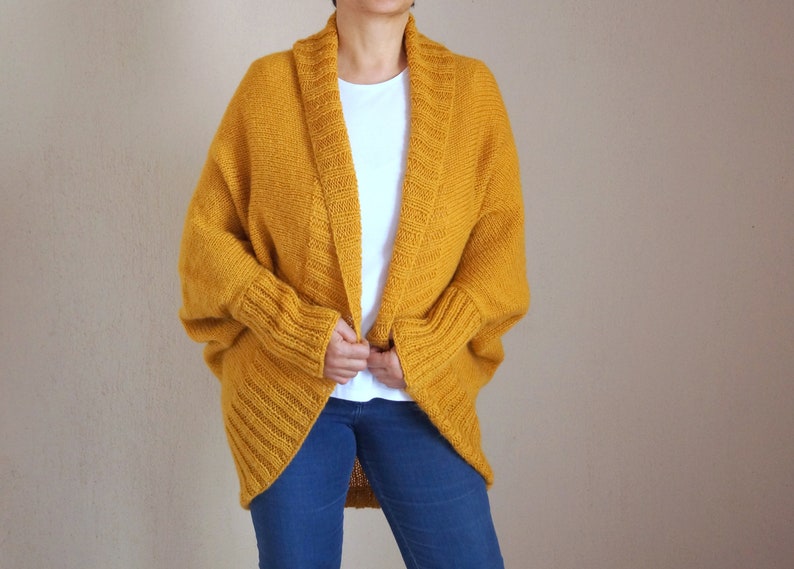 Puede incluir: Un c&aacute;rdigan de punto amarillo mostaza con cuello amplio abierto y mangas largas. El c&aacute;rdigan est&aacute; hecho de un hilo suave y grueso y tiene un corte holgado.