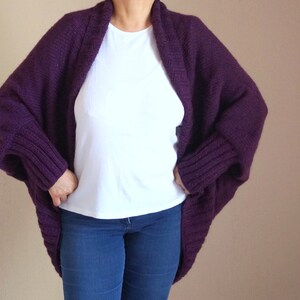 May include: A deep purple, chunky knit cardigan with a wide collar and ribbed cuffs. The open-front sweater is worn over a white t-shirt and blue jeans. The sleeves are long and the cardigan is a loose fit.