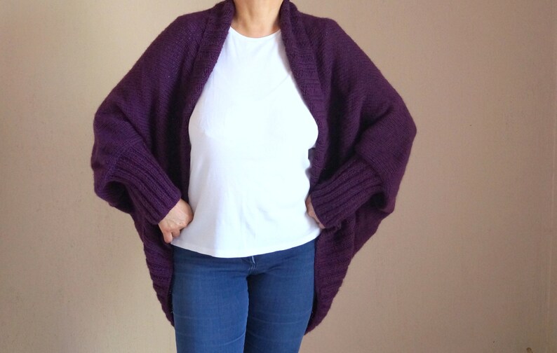 May include: A deep purple, chunky knit cardigan with a wide collar and ribbed cuffs. The open-front sweater is worn over a white t-shirt and blue jeans. The sleeves are long and the cardigan is a loose fit.