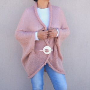 May include: A soft, blush pink knitted cardigan with a long, flowing silhouette. The cardigan is open at the front and has a white circle-shaped clasp.
