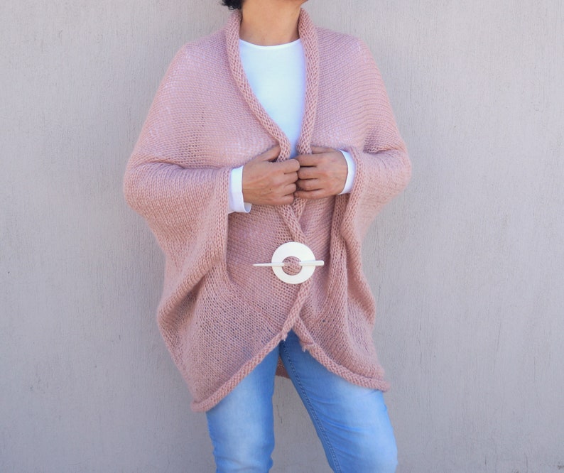 May include: A soft, blush pink knitted cardigan with a long, flowing silhouette. The cardigan is open at the front and has a white circle-shaped clasp.