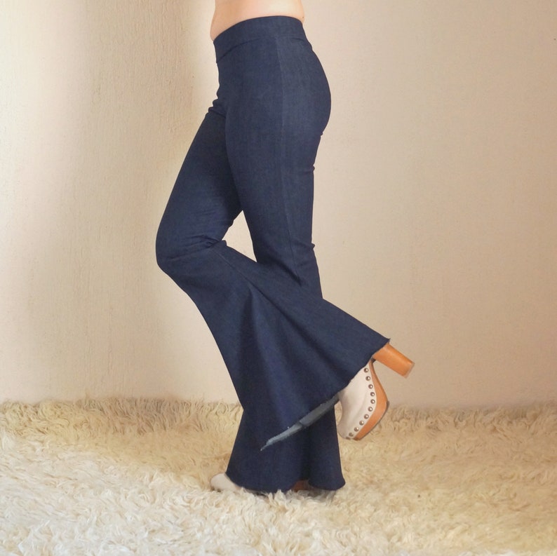 May include: Dark blue denim bell-bottom pants. The pants have a high waist and a wide flare from the knee down. The model is wearing white and tan clogs with a wooden heel.