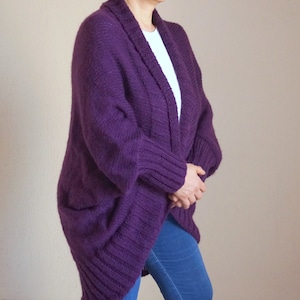May include: A deep purple, open-front cardigan with a shawl collar and two front pockets. The knitted sweater is made of a thick, textured yarn. The person is wearing a white shirt and blue jeans underneath the cardigan.