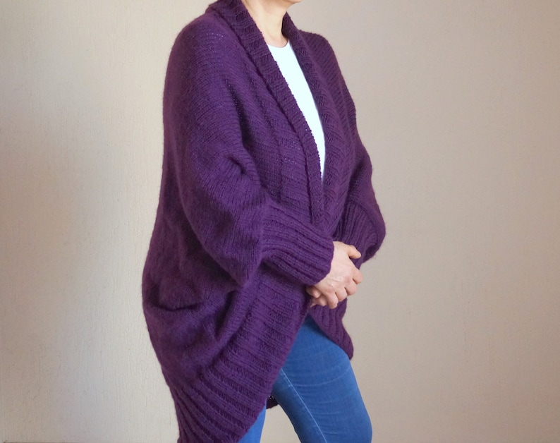 May include: A deep purple, open-front cardigan with a shawl collar and two front pockets. The knitted sweater is made of a thick, textured yarn. The person is wearing a white shirt and blue jeans underneath the cardigan.