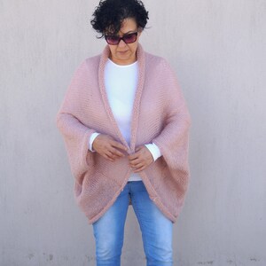 May include: A blush pink knitted cardigan with a wide, open front and long sleeves. The cardigan is made of a soft, lightweight yarn and has a relaxed fit.