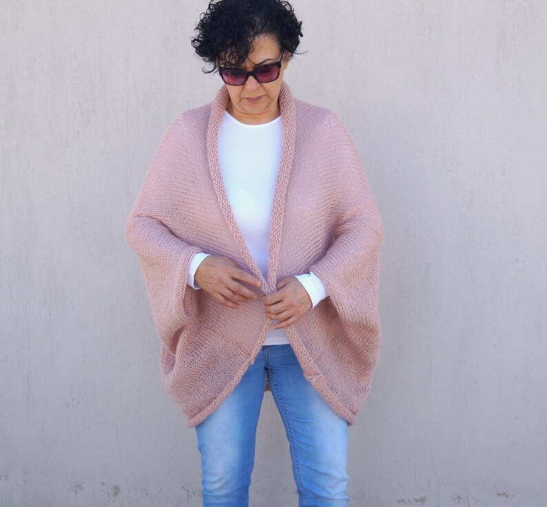 May include: A blush pink knitted cardigan with a wide, open front and long sleeves. The cardigan is made of a soft, lightweight yarn and has a relaxed fit.