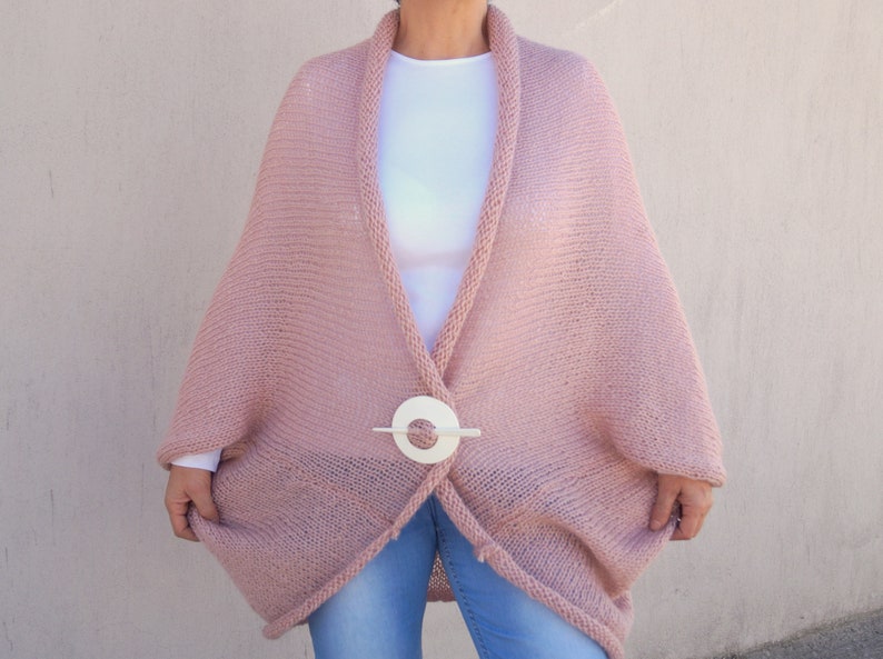 May include: A soft, pink knitted cardigan with a white button closure. The cardigan has a relaxed fit and a long, flowing silhouette.