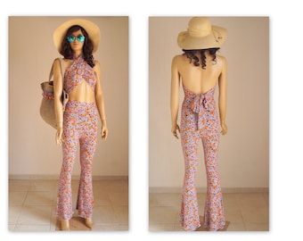 May include: A floral print jumpsuit with a halter top and wide-leg pants. The top has a tie at the back and the pants have a bell-bottom style. The model is wearing a straw hat and sunglasses.